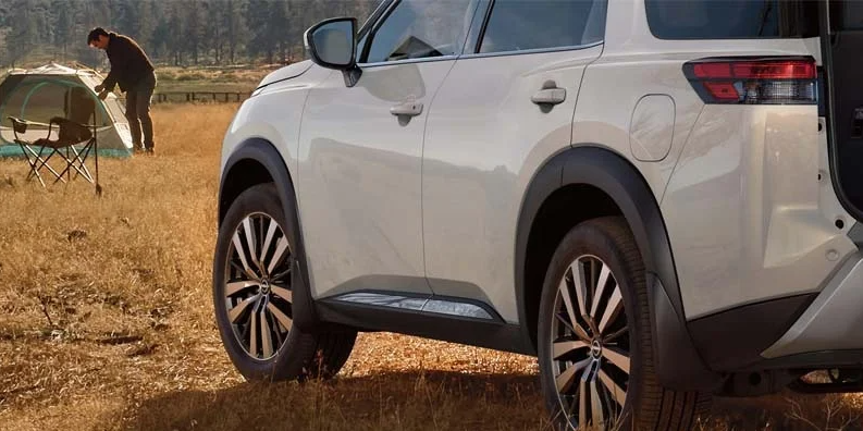A white 2024 Nissan Pathfinder parked near a man setting up a tent for camping.